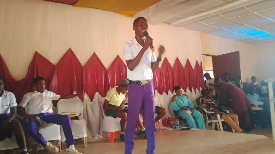 A student in purple trousers speaks into a microphone on stage, 2025