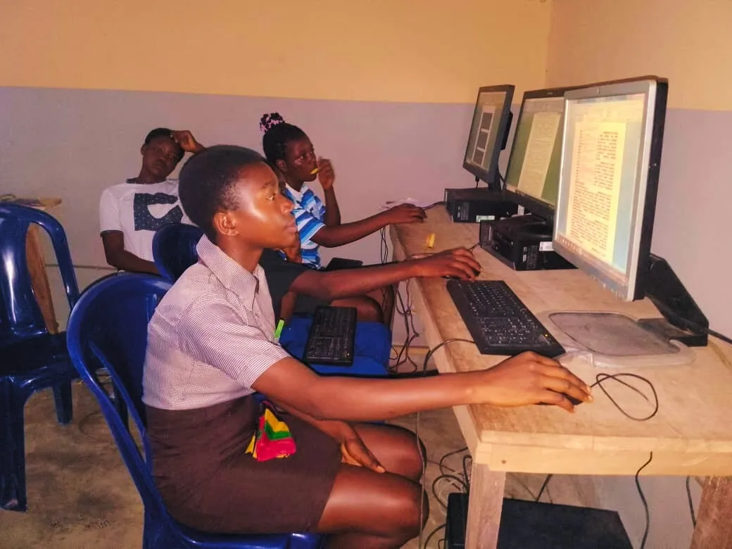 A girl in school uniform works at a computer while classmates look on