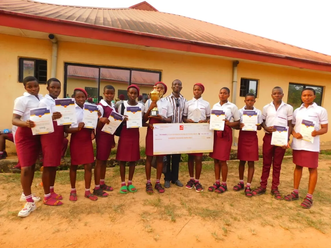 Students in maroon uniforms hold certificates, a trophy and a cheque outside the hall